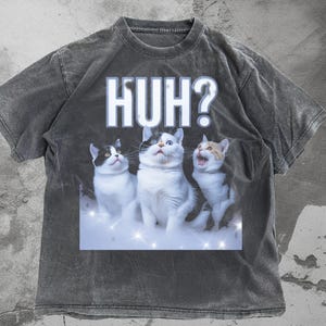 May include: Dark gray t-shirt featuring a graphic of three cats with the word "HUH?" above them. The cats are white with black and orange markings, set against a sparkling background. The shirt has a vintage, washed look.