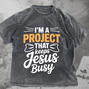 Puede incluir: Una camiseta gris oscuro con el texto "I'M A PROJECT THAT keeps Jesus Busy" en blanco y naranja. La camiseta tiene un aspecto vintage y desgastado.