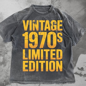 May include: A gray t-shirt with the words "VINTAGE 1970'S LIMITED EDITION" in a yellow, distressed font. The shirt has a vintage, worn look, with a crew neck and short sleeves. The text is stacked vertically.