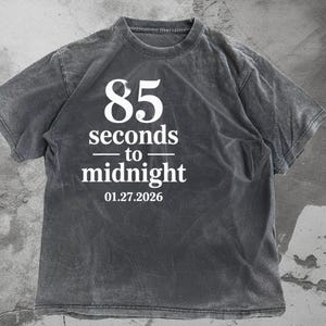May include: A dark gray t-shirt with the text "85 seconds to midnight" in white, along with the date 01.27.2026. The shirt has a vintage, distressed look.