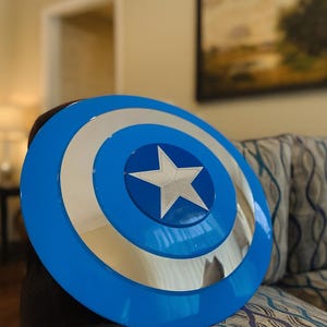 May include: A blue and silver Captain America shield. The shield has a blue outer ring, a silver inner ring, and a silver star in the center. The shield is round and appears to be made of metal.