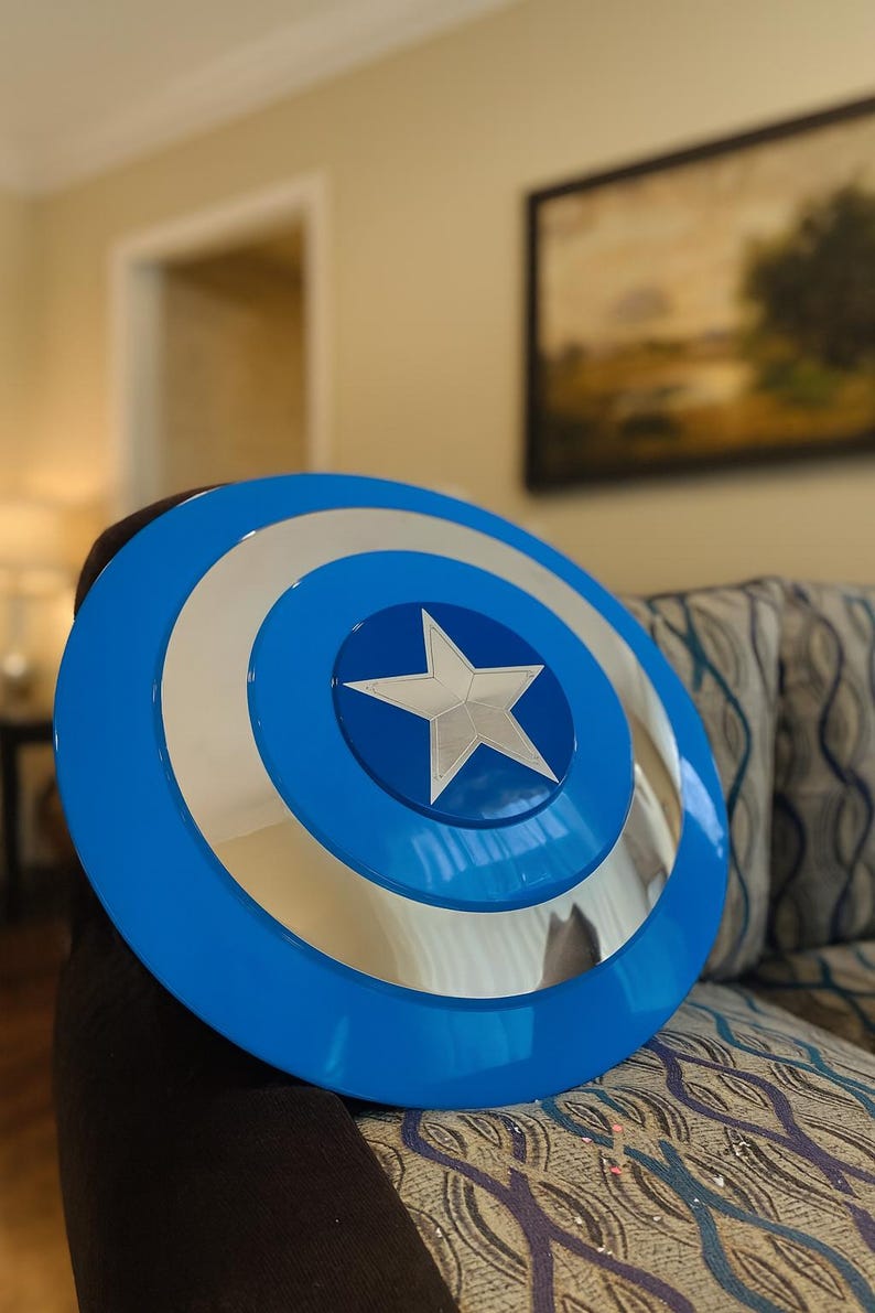 May include: A blue and silver Captain America shield. The shield has a blue outer ring, a silver inner ring, and a silver star in the center. The shield is round and appears to be made of metal.