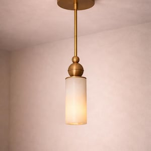 May include: A brass pendant light with a cylindrical, frosted glass shade. The light hangs from a brass rod and a circular ceiling mount. The fixture emits a warm glow. The height is approximately 30 cm.