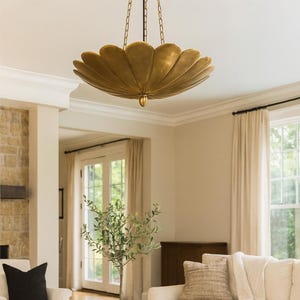 May include: A decorative, gold-toned chandelier with a petal design hangs from the ceiling. The light fitting is suspended by chains. The room has a neutral colour scheme, with white walls, a white sofa, and a wooden coffee table.