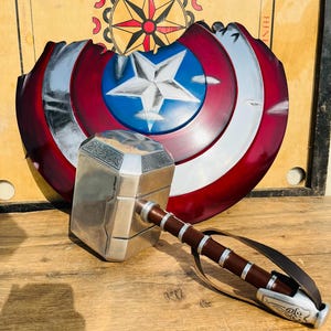 May include: A silver and brown hammer with a leather strap and a Captain America shield in the background. The shield is red, white, and blue with a silver star in the center. The hammer has intricate detailing.