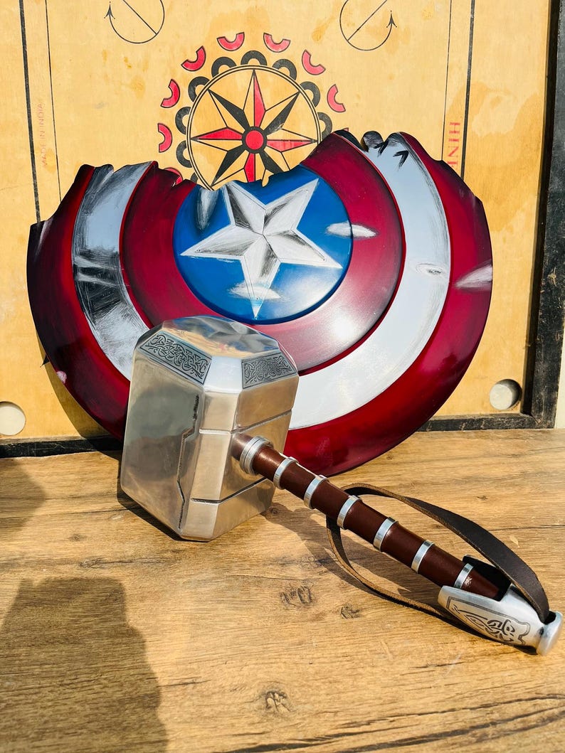 May include: A silver and brown hammer with a leather strap and a Captain America shield in the background. The shield is red, white, and blue with a silver star in the center. The hammer has intricate detailing.
