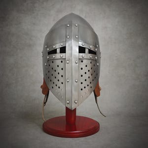 Può includere: Un elmo medievale color argento con la parte superiore arrotondata e strisce di metallo rivettate. L'elmo presenta fessure per gli occhi e numerosi piccoli fori di ventilazione. È appoggiato su un supporto di legno rosso con cinghie di cuoio.