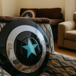 May include: A black and silver shield with a blue star emblem, resting on a patterned armchair. The shield has a circular design with a silver ring and a central blue star. The armchair has a brown cushion and a patterned fabric.