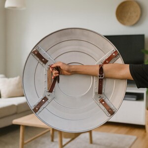 May include: A silver-colored, round shield with brown leather straps and rivets. The shield has a central circular design and is held by a person's arm. The shield is a prop or costume piece.