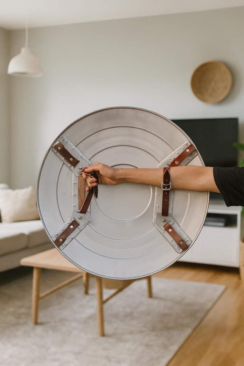 May include: A silver-colored, round shield with brown leather straps and rivets. The shield has a central circular design and is held by a person's arm. The shield is a prop or costume piece.