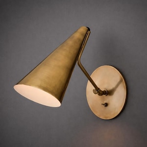 May include: A brass wall sconce featuring a conical shade and a circular backplate. The shade is angled downwards, and the backplate includes a small switch. The fixture has a warm gold finish.