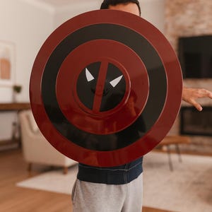 May include: A round, red and black shield with a Deadpool design. The shield has concentric circles in red and black, with a white eye and mouth design in the center. The shield is held in front of a person.