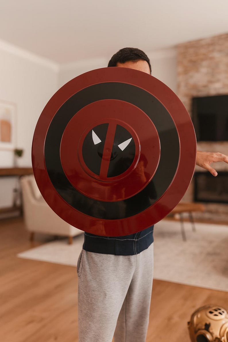 May include: A round, red and black shield with a Deadpool design. The shield has concentric circles in red and black, with a white eye and mouth design in the center. The shield is held in front of a person.