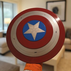 May include: A red, white, and blue Captain America shield. The shield is round with a silver band and a blue circle in the center, featuring a white star. The shield is held up in front of a blurred background.