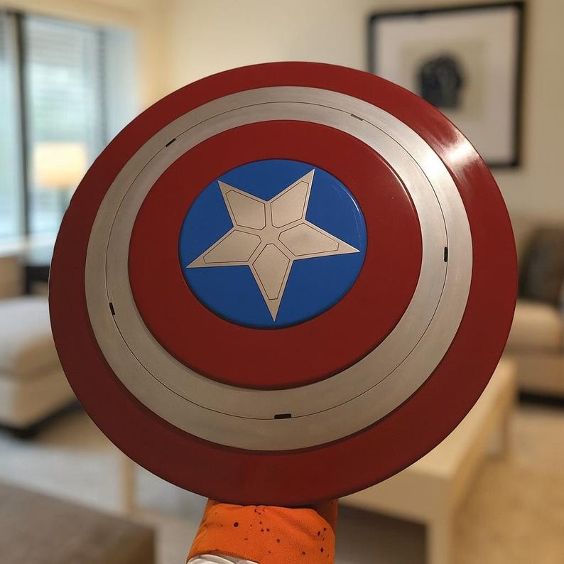 May include: A red, white, and blue Captain America shield. The shield is round with a silver band and a blue circle in the center, featuring a white star. The shield is held up in front of a blurred background.