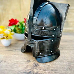 Peut inclure: Casque en métal noir avec des détails rivetés et une visière rectangulaire. Le casque a un dessus arrondi et une protection de cou. Le design suggère un thème historique ou fantastique, peut-être pour l'affichage ou le costume.
