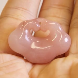 以下が含まれることがあります： 中心にハート型の切り抜きがある、薄いピンク色の花型のペンダント。 ペンダントは滑らかで磨かれており、半透明です。 開いた手に持たれています。