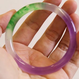 May include: A multi-colored jade bangle bracelet. The bracelet transitions from green to white to purple. The smooth, polished surface reflects light, highlighting the natural variations in color. The bracelet is held in a hand against a neutral background.