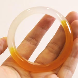 Puede incluir: Un brazalete circular translúcido con un degradado de colores. El brazalete pasa de transparente a un tono naranja vibrante. La joya se sostiene en una mano sobre un fondo neutro. Este accesorio es una adición elegante a cualquier colección de joyas.