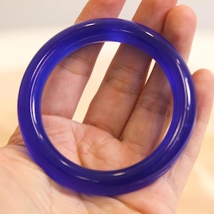 May include: A translucent, deep blue bangle bracelet. The circular bracelet is held in a hand, showcasing its smooth, polished surface. The color is a vibrant, saturated blue, with a glossy finish. The bangle is a classic jewelry piece.