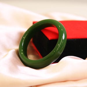 Puede incluir: Un brazalete de jade verde oliva brillante. El brazalete circular liso se muestra sobre una tela color crema, con una caja de joyería roja y negra en el fondo. Una pieza de joyería clásica.