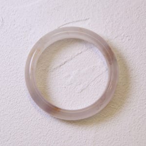 Op de afbeelding: Een doorschijnende, lavendelkleurige armband. Het gladde, gepolijste oppervlak van de armband heeft subtiele kleurvariaties, met hints van bruin en wit. De cirkelvorm is eenvoudig en elegant, geschikt voor dagelijks gebruik.