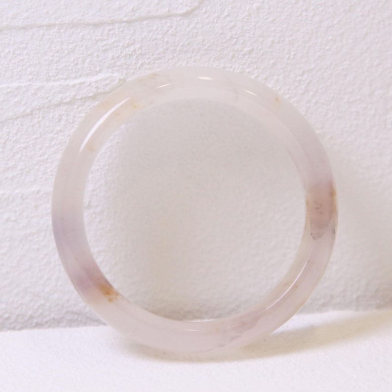 Op de afbeelding: Een doorschijnende, lichtpaarse armband. De gladde, cirkelvormige armband heeft subtiele kleurvariaties, met hints van perzik en wit. De sieraden zijn geplaatst tegen een getextureerde witte achtergrond, wat de delicate uitstraling benadrukt.