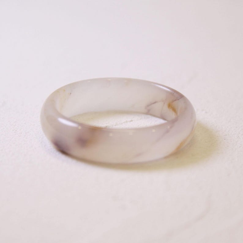 Op de afbeelding: Een doorschijnende, lichtpaarse ring met subtiele bruine en witte marmering. De gladde, afgeronde band is een eenvoudig, elegant ontwerp. Deze ring is een sieraad.