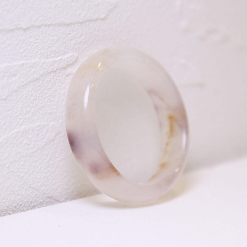 Op de afbeelding: Een doorschijnende ring met een gepolijste afwerking. De ring is voornamelijk wit met subtiele paarse en goudkleurige insluitsels. Het gladde oppervlak reflecteert licht, waardoor de kleur- en textuurvariaties worden benadrukt. Een eenvoudig, elegant sieraad.