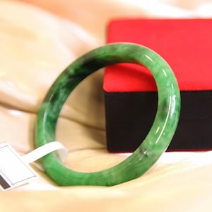 May include: A jade bangle bracelet with a smooth, polished surface. The bracelet is a vibrant green colour with darker green marbling. A white tag is attached to the bracelet. A red and black box is in the background.