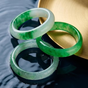 May include: Three jade bangles in varying shades of green and white rest on a dark, reflective surface. The bangles are smooth and polished, with natural variations in color. A gold-colored tray is in the background.