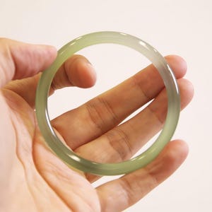 Puede incluir: Un brazalete de jade verde pálido. El brazalete circular se sostiene en una mano. El jade tiene un sutil degradado de color, con un tono ligeramente más oscuro hacia el centro. El fondo es blanco.