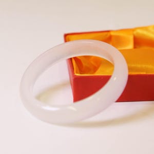 Puede incluir: Un brazalete de jade translúcido y pálido. El brazalete liso y redondeado se muestra frente a una caja roja con un forro de satén amarillo. El brazalete tiene un brillo suave y nacarado.