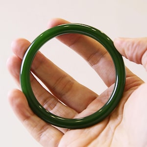 May include: A dark green jade bangle. The polished surface reflects light, showing the rich colour. The circular design is simple and elegant. The bangle is held in a hand. The diameter is approximately 6.5 cm.