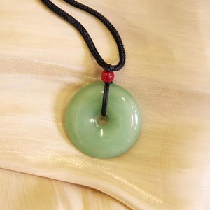 Op de afbeelding: Een jade hangerketting met een cirkelvormige, lichtgroene steen met een centraal gat. De hanger hangt aan een zwart koord, geaccentueerd door een kleine rode kraal. De ketting wordt gepresenteerd tegen een zachte, crèmekleurige achtergrond.