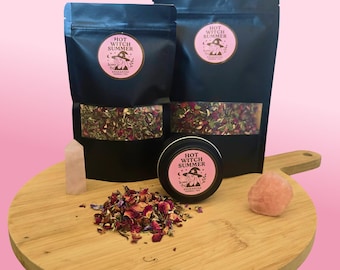 May include: An arrangement of products on a wooden cutting board against a pink background. The items include two black pouches and a small tin, all labeled "Hot Witch Summer." Also present are a pile of dried flowers, a crystal, and a pink stone.