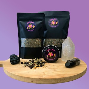 May include: Black pouches and a tin of "GIRL, BYE" enchanted herbal tea, with a witch illustration on the label. The tea is displayed on a wooden board with crystals and loose tea. The background is a solid purple.