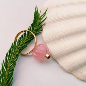 May include: A gold ring rests on a green sprig, next to a pink, carved glass bead with a gold-colored metal cap. A large, white seashell provides a textured backdrop.