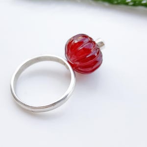 May include: A silver ring featuring a vibrant red, carved glass bead. The bead has a rounded shape with a textured surface. The ring is set against a white background, highlighting the jewelry's design.