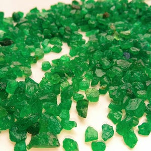May include: A close-up image displays a collection of raw emeralds. The gemstones are a vivid, translucent green, with diverse shapes and sizes. The uncut emeralds are scattered on a white surface, creating a visually appealing arrangement.