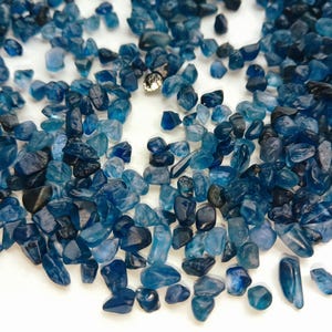 Puede incluir: Una colección de pequeñas gemas azules de forma irregular. Las piedras varían en tonos de azul, de claro a oscuro, y están dispersas sobre una superficie blanca. También hay un pequeño objeto brillante de color plateado.