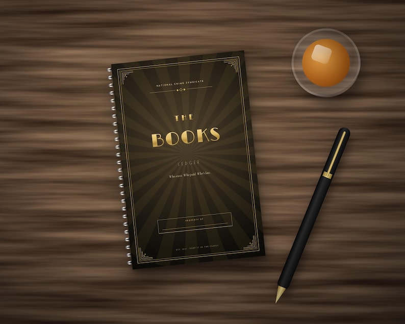 May include: A spiral-bound notebook with a dark brown cover featuring a gold sunburst pattern and the text THE BOOKS LEDGER. A black pen with gold accents and a small glass dish containing a round orange object are positioned nearby.
