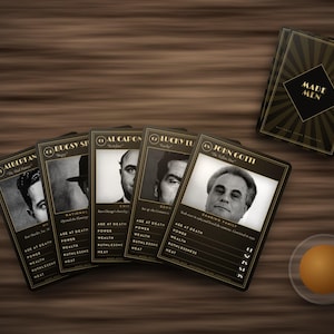 May include: A set of Made Men playing cards featuring black and gold borders with portraits of historical figures. The cards display statistics for age at death, power, wealth, ruthlessness, and heat. A small orange sphere sits nearby.