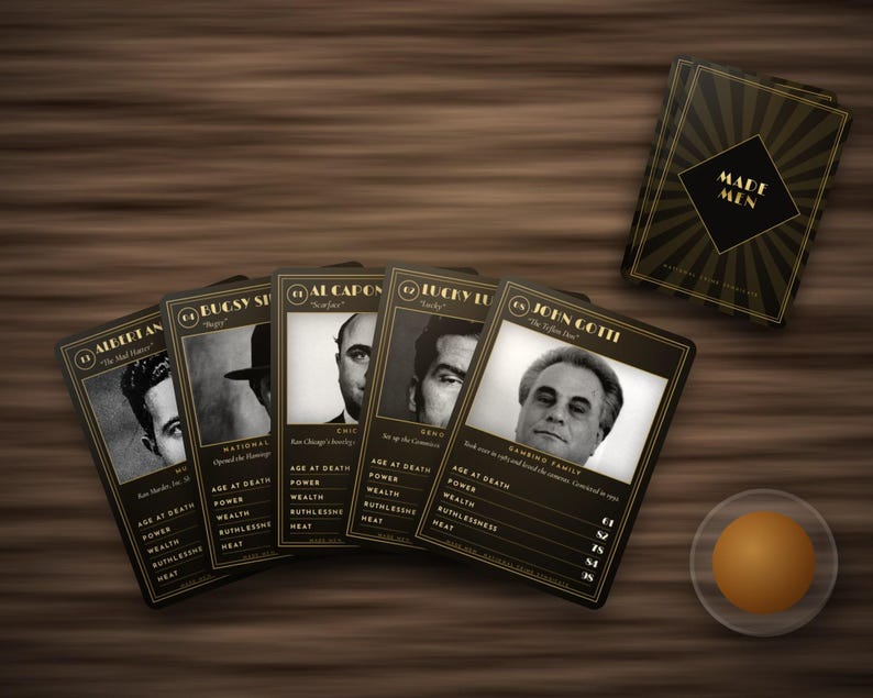 May include: A set of Made Men playing cards featuring black and gold borders with portraits of historical figures. The cards display statistics for age at death, power, wealth, ruthlessness, and heat. A small orange sphere sits nearby.