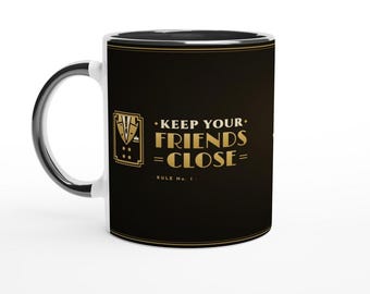May include: A black ceramic mug with a white interior and handle. The exterior features a gold graphic of a tuxedo jacket and the text: KEEP YOUR FRIENDS CLOSE = RULE No. 1. The design is framed by thin gold lines at the top and bottom edges.