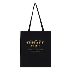 May include: A black tote bag with two long shoulder straps. The front features gold text that reads MADE IN THE CHICAGO OUTFIT, followed by five gold stars and the text CHICAGO ILLINOIS, all centered on the bag's surface.