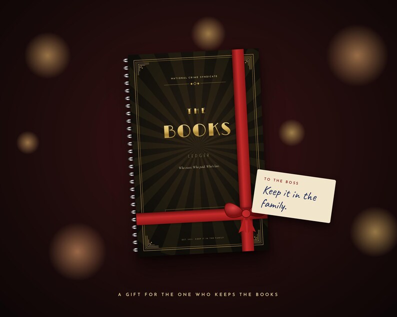 May include: A black spiral-bound ledger titled THE BOOKS with gold lettering and a sunburst pattern, wrapped in a red ribbon with a gift tag that reads TO THE BOSS Keep it in the family. The bottom text reads A GIFT FOR THE ONE WHO KEEPS THE BOOKS.