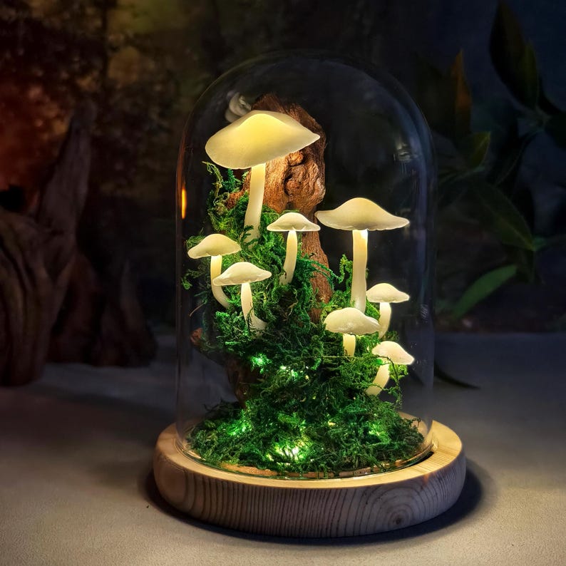 May include: A decorative glass cloche featuring illuminated mushroom sculptures. The mushrooms are white with yellow lighting, set amidst green moss and a piece of wood. The cloche sits on a round wooden base, creating a whimsical, nature-inspired display.