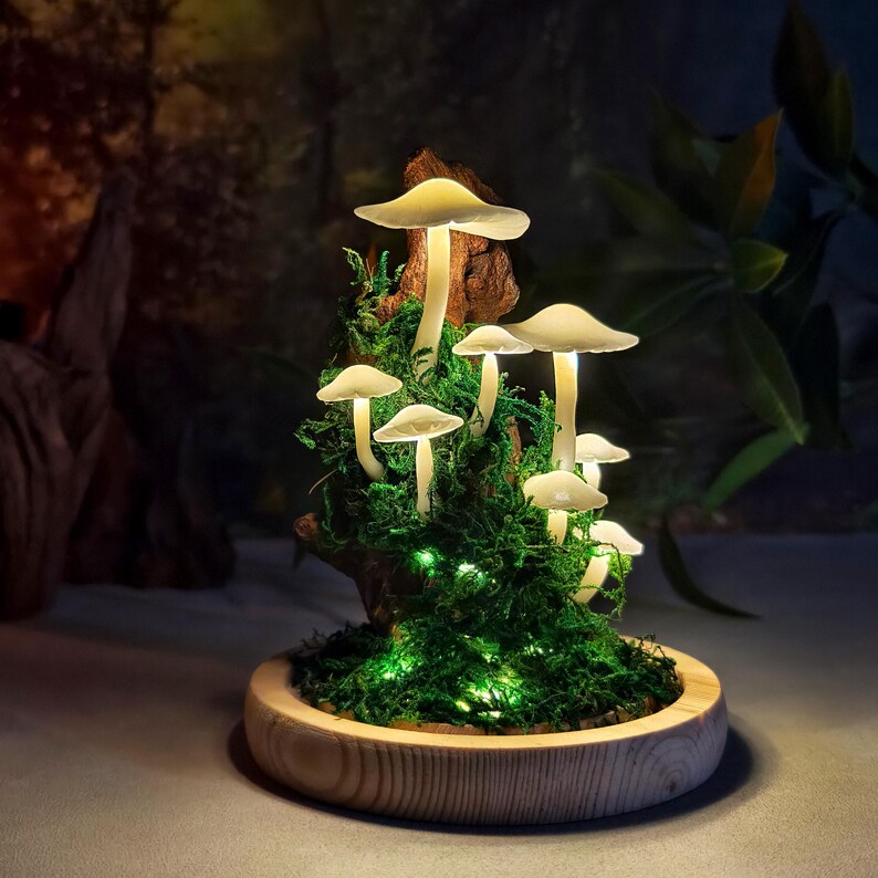 May include: A decorative mushroom lamp with glowing white mushroom caps and stems. The mushrooms are set in green moss and a wooden base. The lamp is illuminated with small green lights, creating a whimsical, fairy-tale aesthetic.