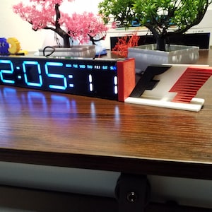 May include: A black digital clock displaying 12:05 with blue numerals, the day of the week, and the date. A red and white F1 logo is on the right. The clock is on a brown wooden surface.